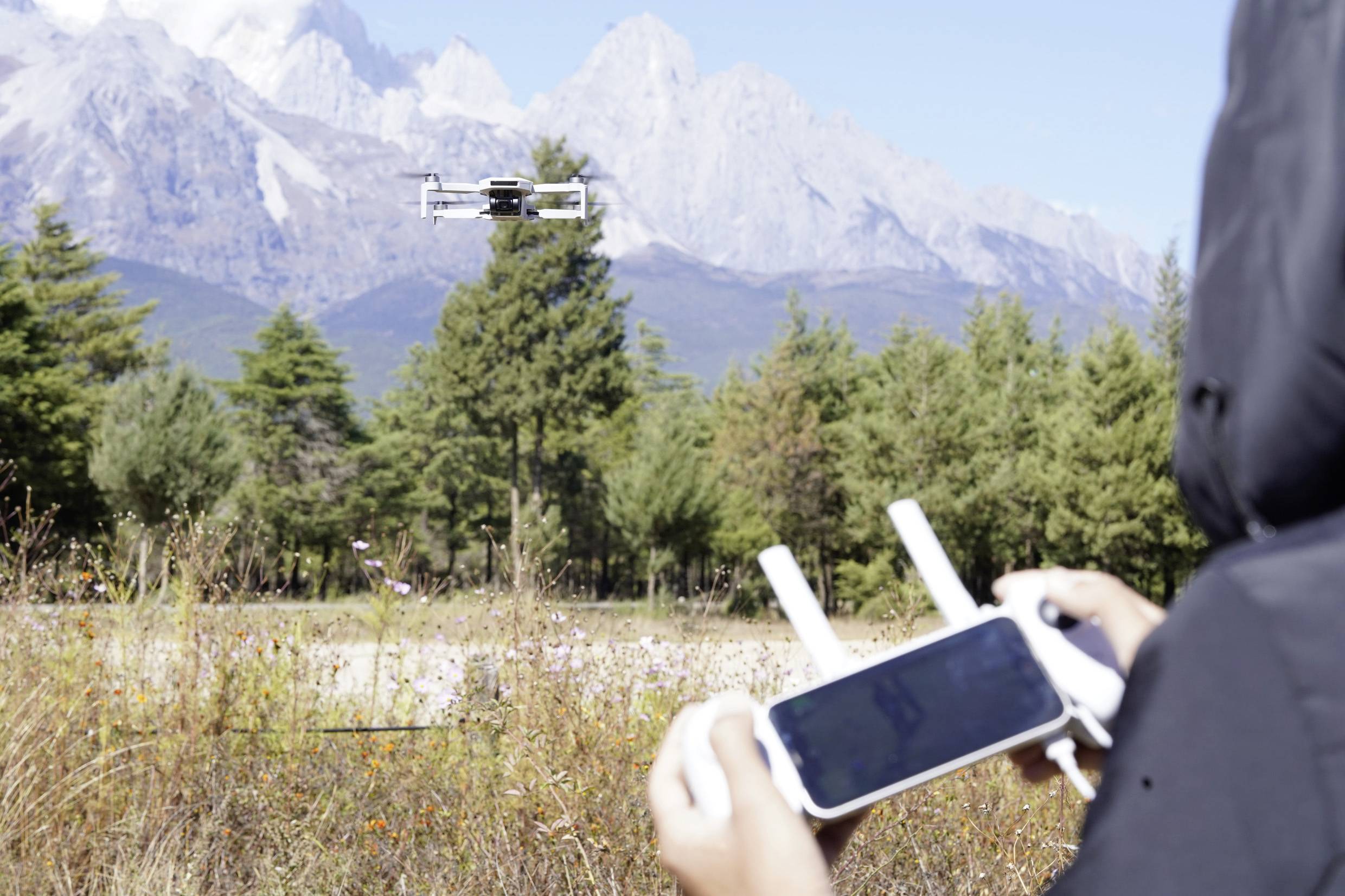 A person is controlling a drone outdoors using a remote control. The drone is hovering in front of a mountainous woodland landscape.