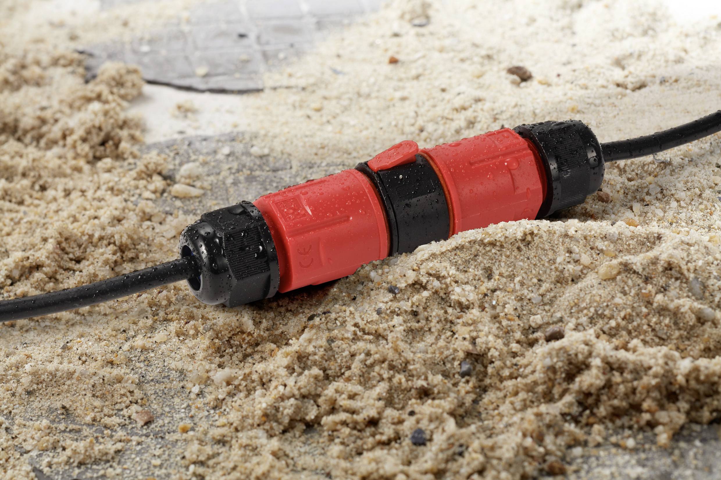 A waterproof connector lies on sandy ground. It is red and black, with visible moisture on its surface.