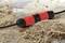 A waterproof connector lies on sandy ground. It is red and black, with visible moisture on its surface.