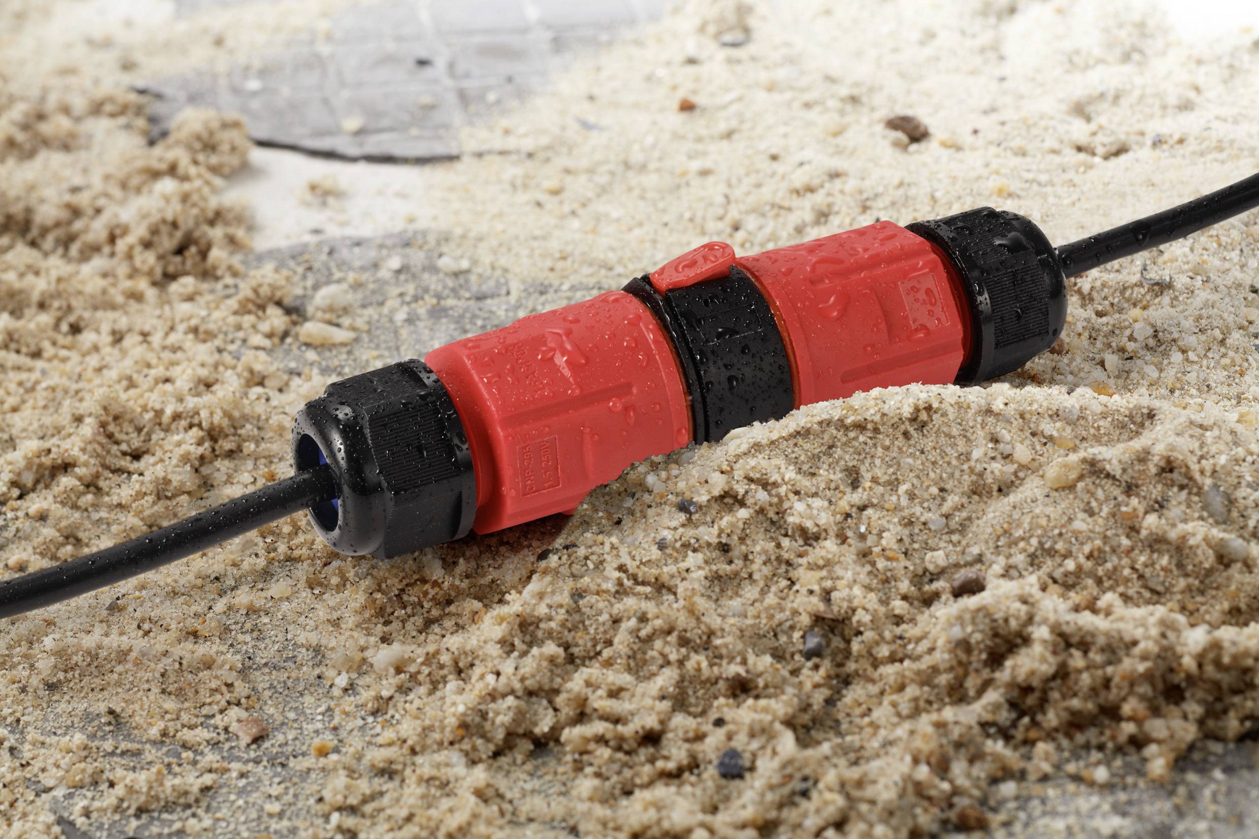 Black cable with red, waterproof connector lies partially in sand. The connector shows water droplets and sand grains.