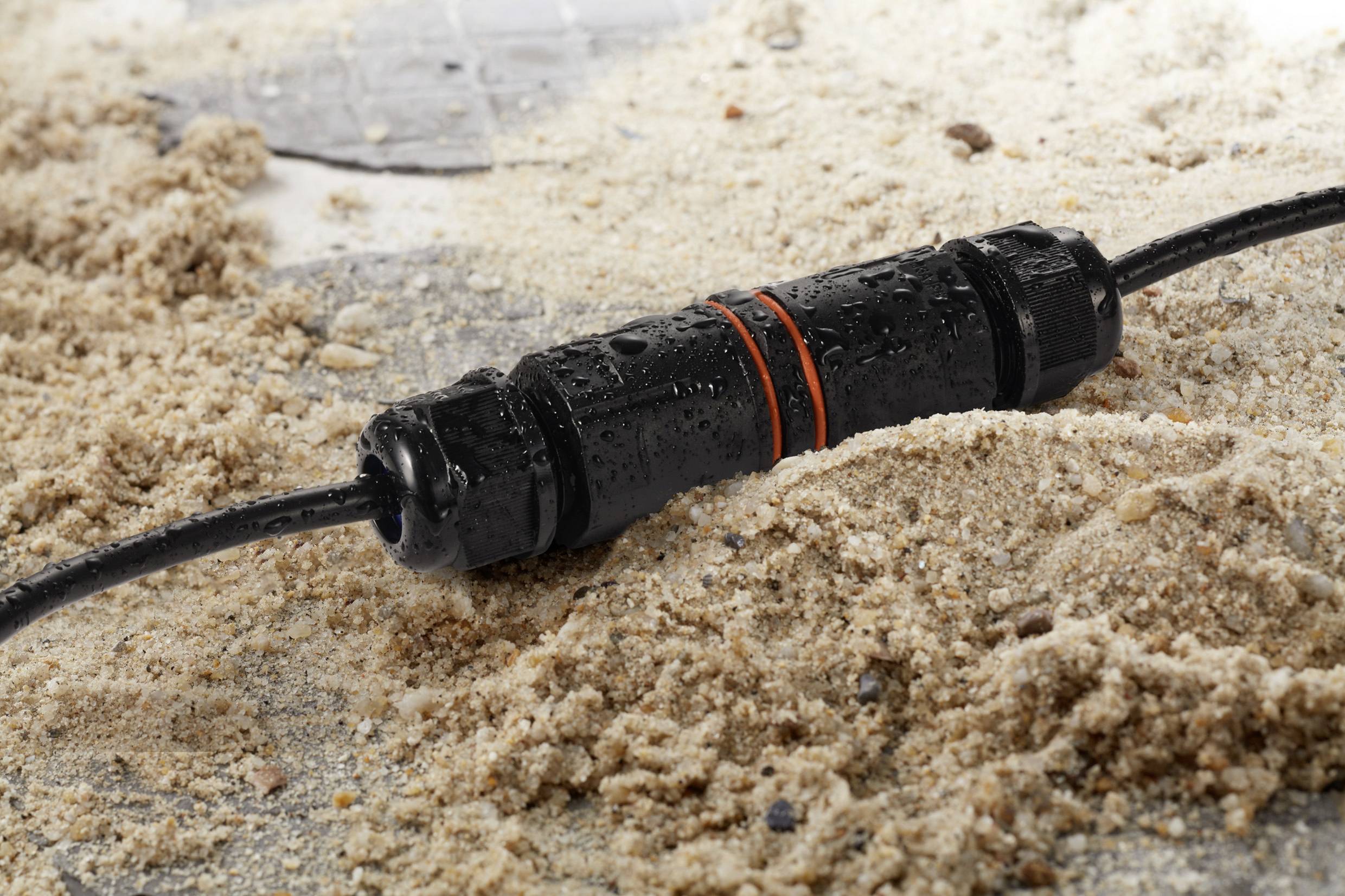 A waterproof cable connector lies in the sand. The connector is black with an orange marking and has water droplets on it.