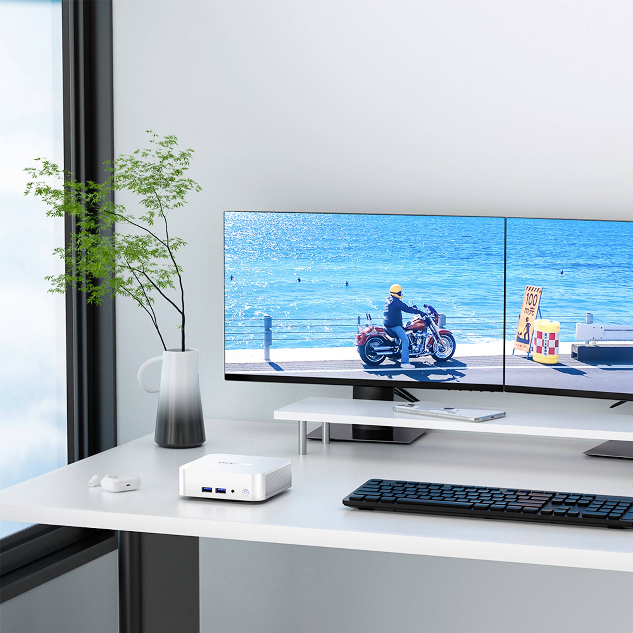 Desk with monitor, keyboard and PC. On the screen an image of a coastal road with a motorcyclist. Plant on the left.
