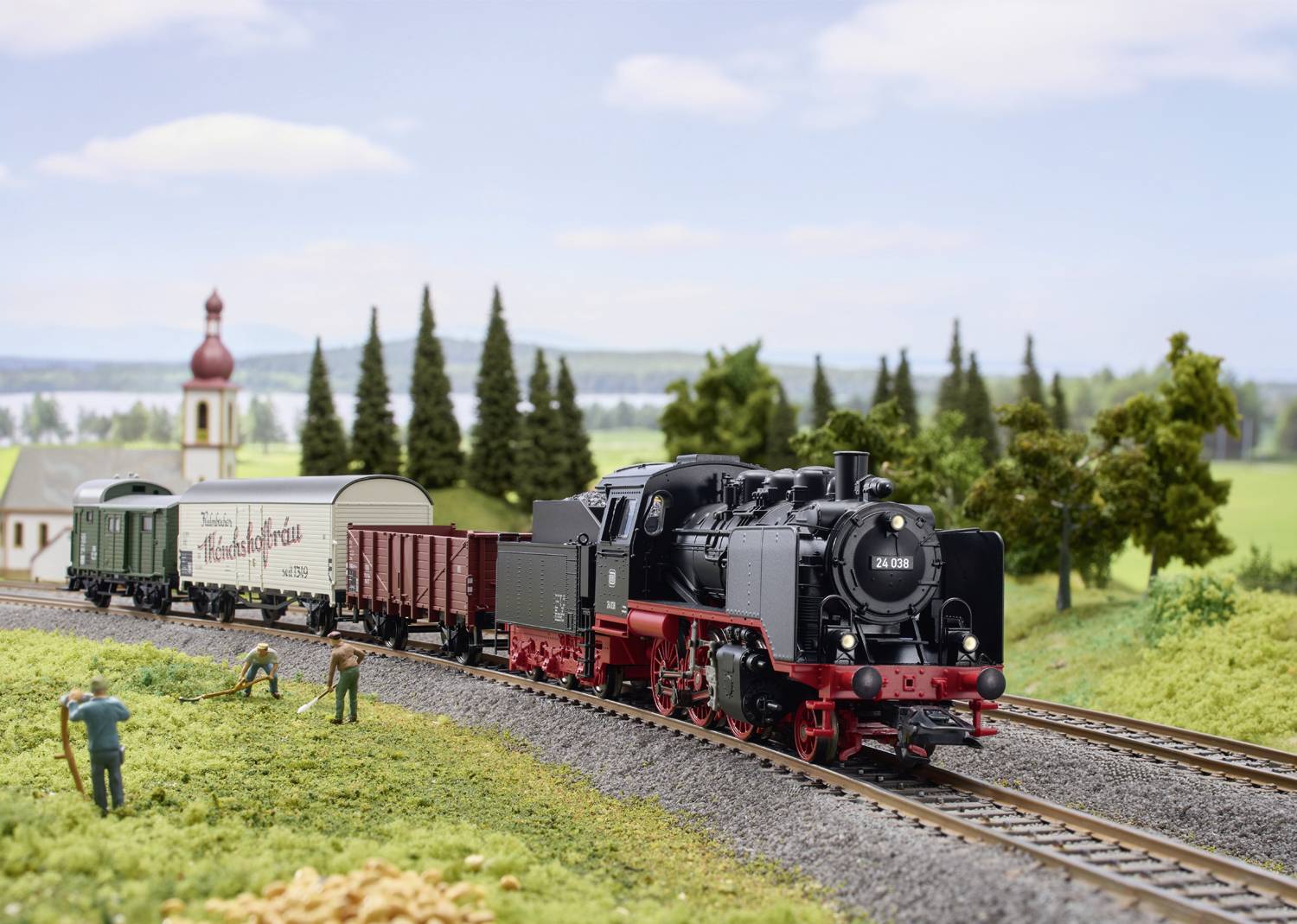 A model train travels through a rural landscape, passing a small church and working figurines in a field.