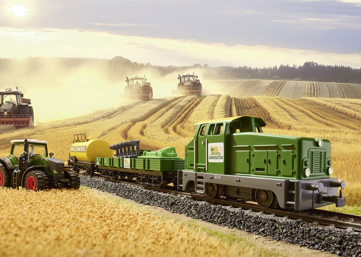 A toy train is transporting agricultural machinery on a model railway track through a landscape with harvesting equipment in operation.