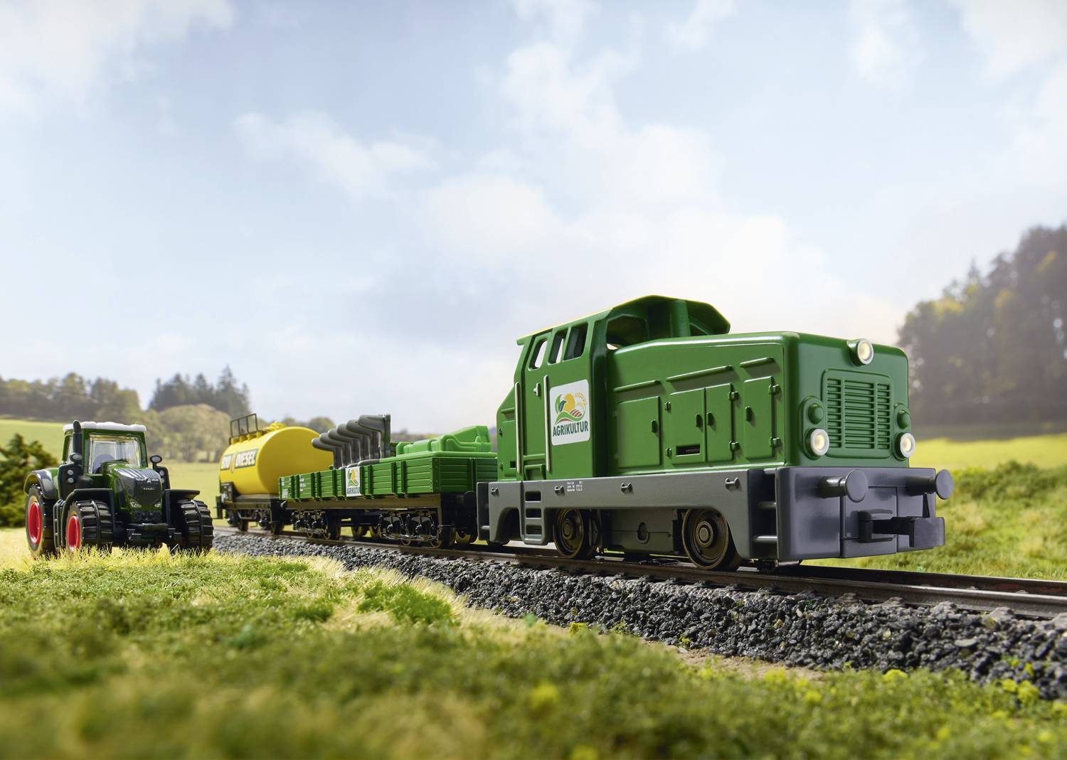 A green train travels along a rural route, accompanied by a tractor in a field. Both vehicles are loaded with agricultural equipment.