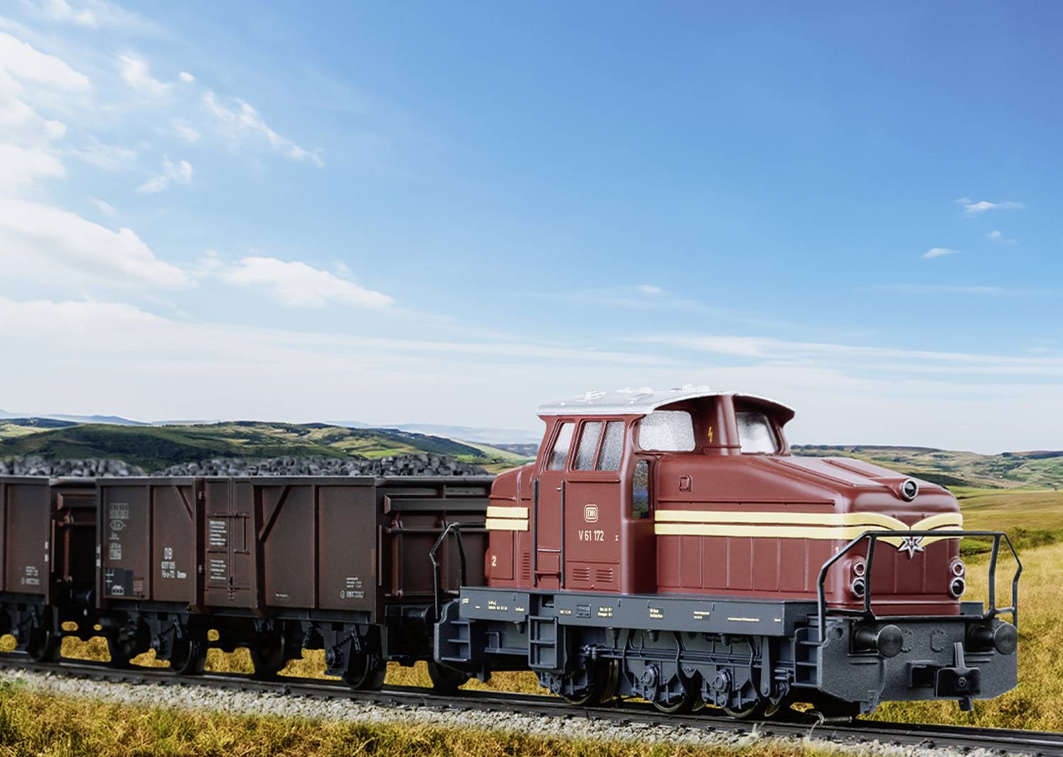 A red locomotive pulls brown freight wagons along a railway line through a rural landscape with green hills beneath a blue sky.
