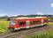 Red regional train on a rural railway line, surrounded by green meadows and hills under a blue sky.