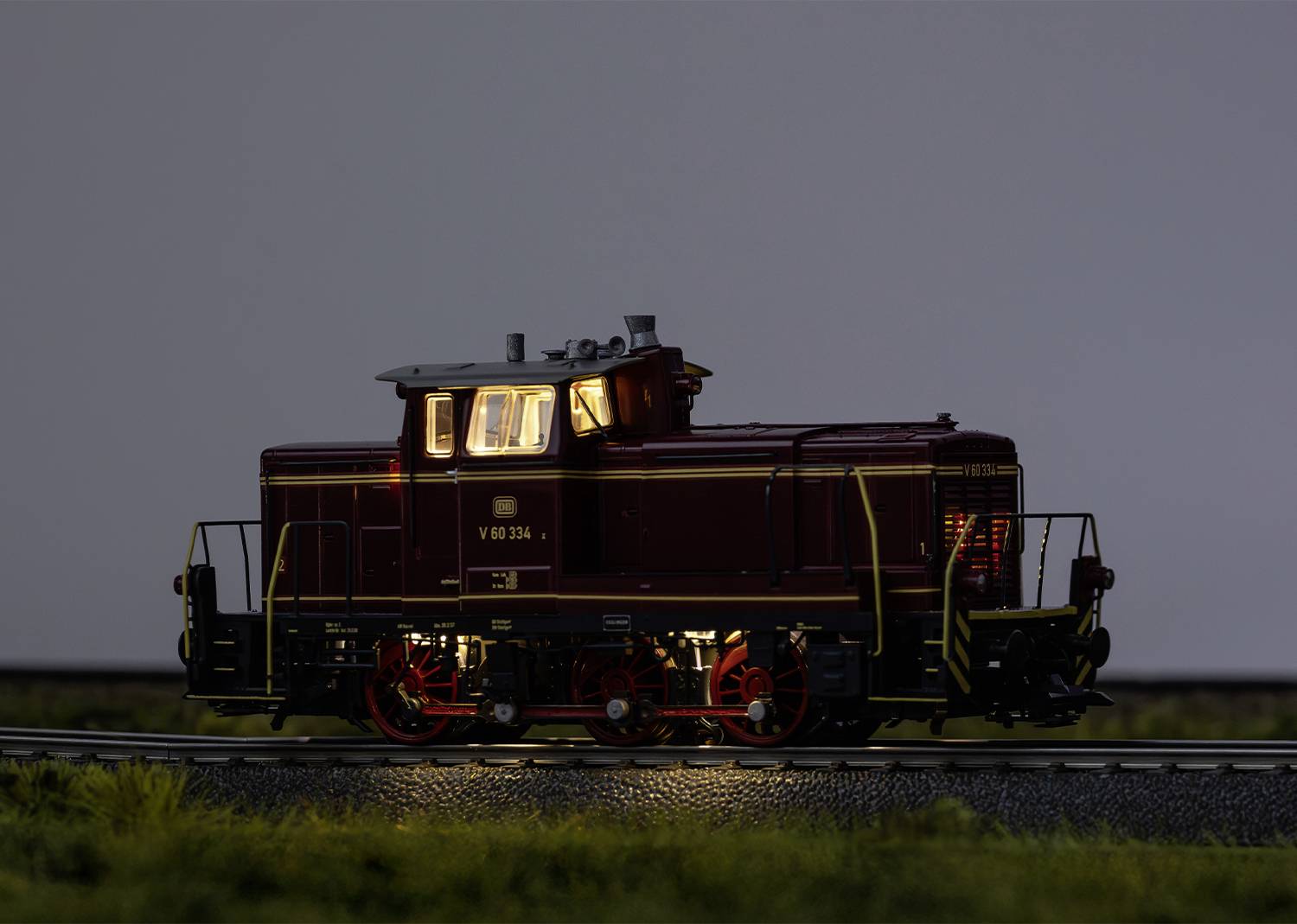 A model train in red colour travels along a railway section. The train's lights are on, and it moves through a scenic landscape.