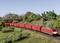 A red goods train travels through a green landscape beneath an iron bridge. Model railway scene with trees and detailed surroundings.