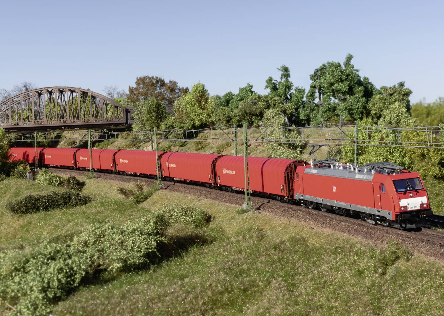 A red freight train is travelling through a green landscape. In the background, a railway bridge and wooded terrain can be seen.