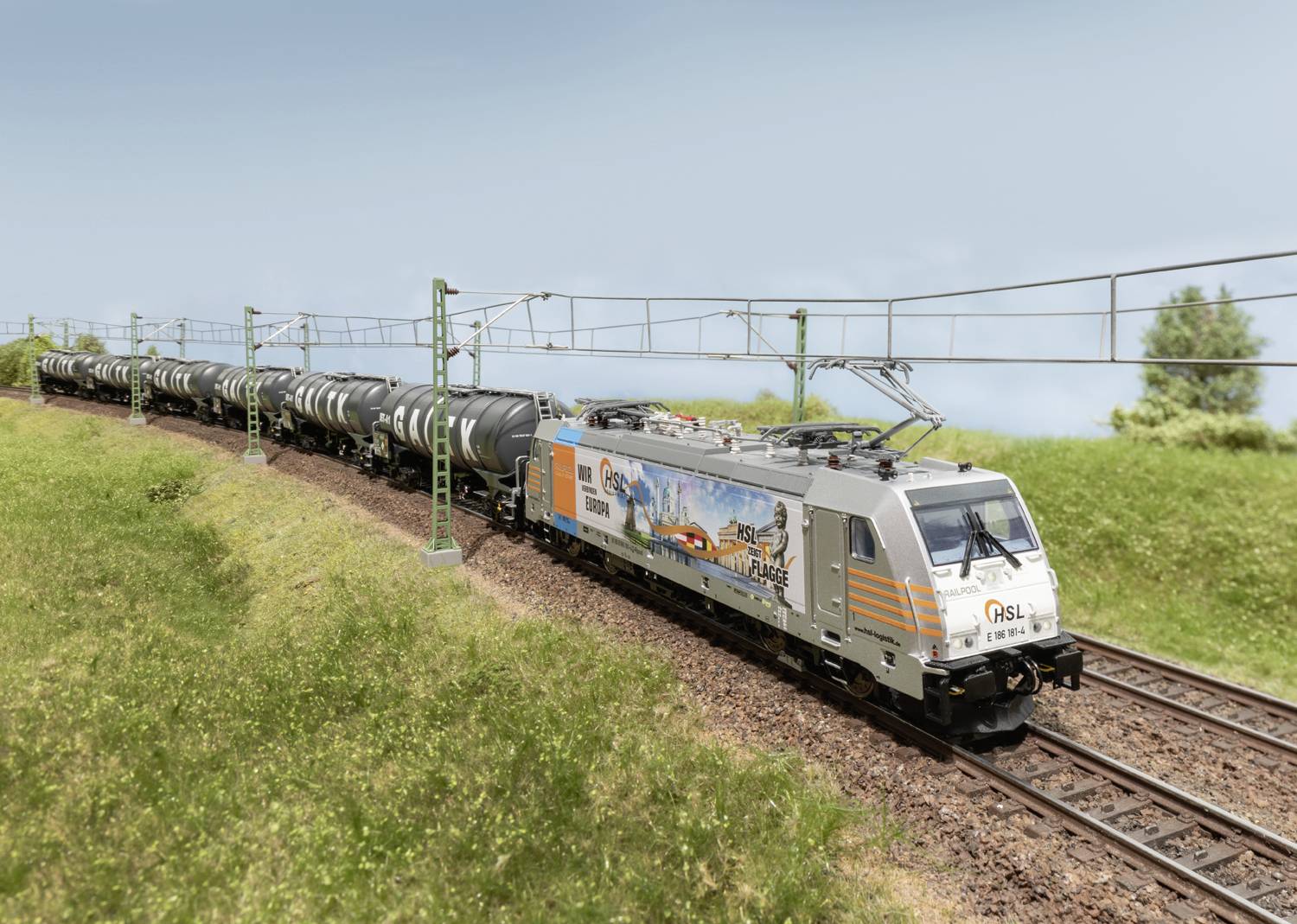 A freight train with tanker wagons travels along a rural railway line. A colourful advertising graphic is visible on the locomotive.