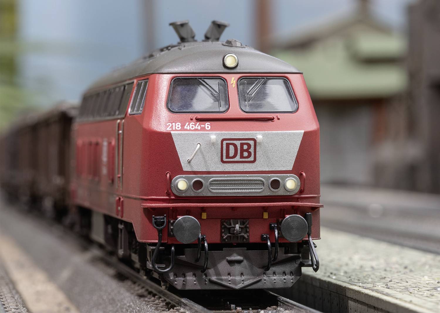 Red locomotive with DB logo travelling on railway track. It carries the number 218 464-6. Background shows blurred railway station.