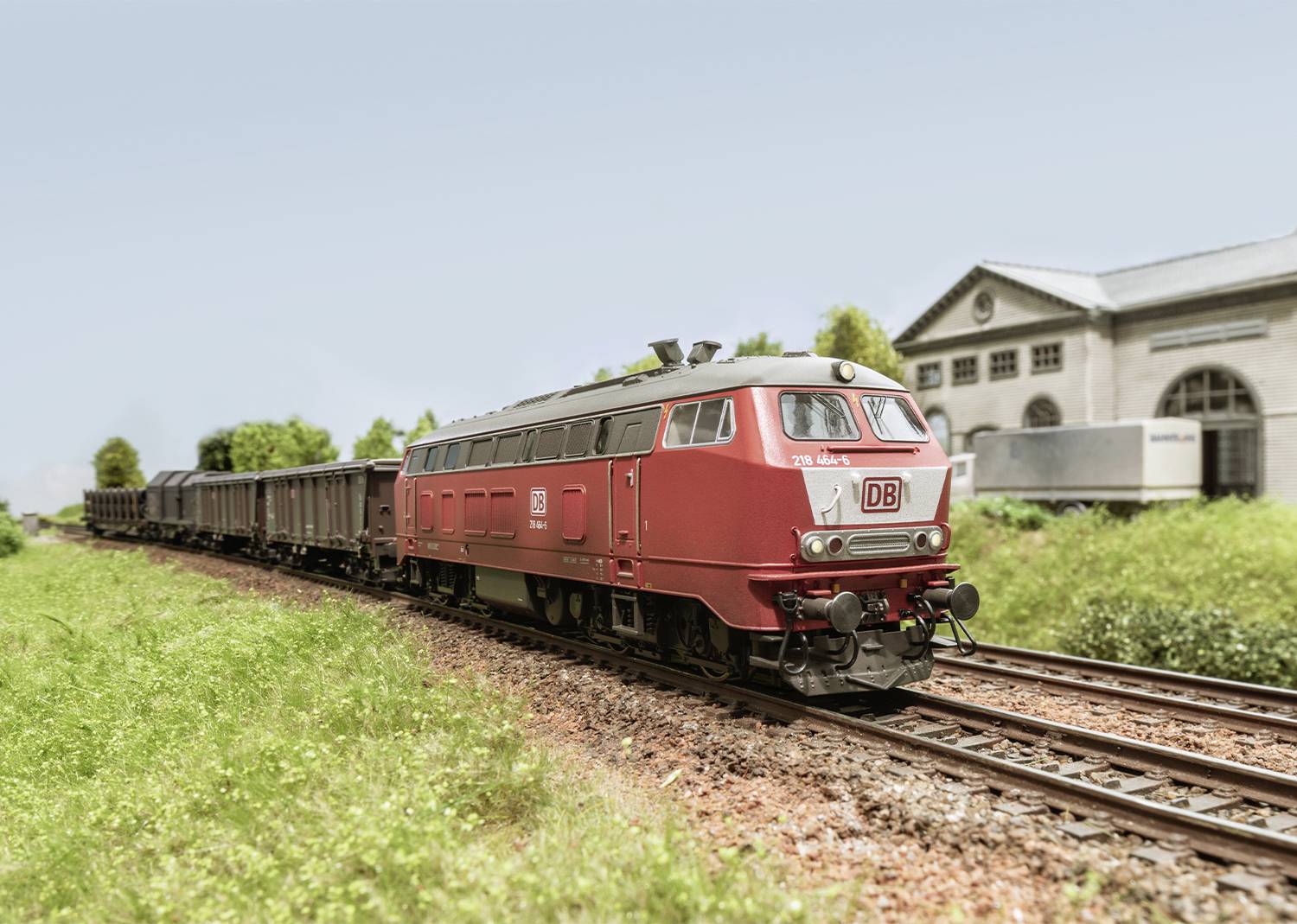 A red freight train of Deutsche Bahn travels in daylight alongside a green meadow, with a railway station building visible in the background.