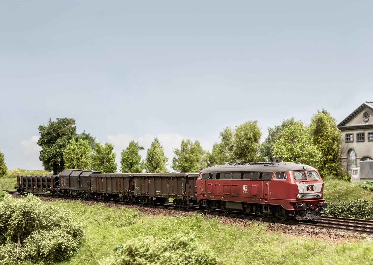 A red goods train travels along a rural route, surrounded by trees and meadows. In the background, a grey building stands.