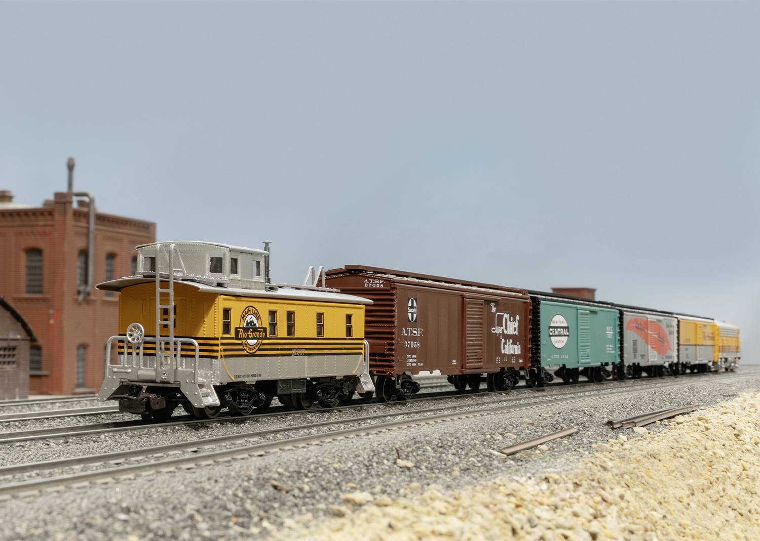 Model railway with coloured goods wagons on tracks, brown brick building in the background, clear sky.