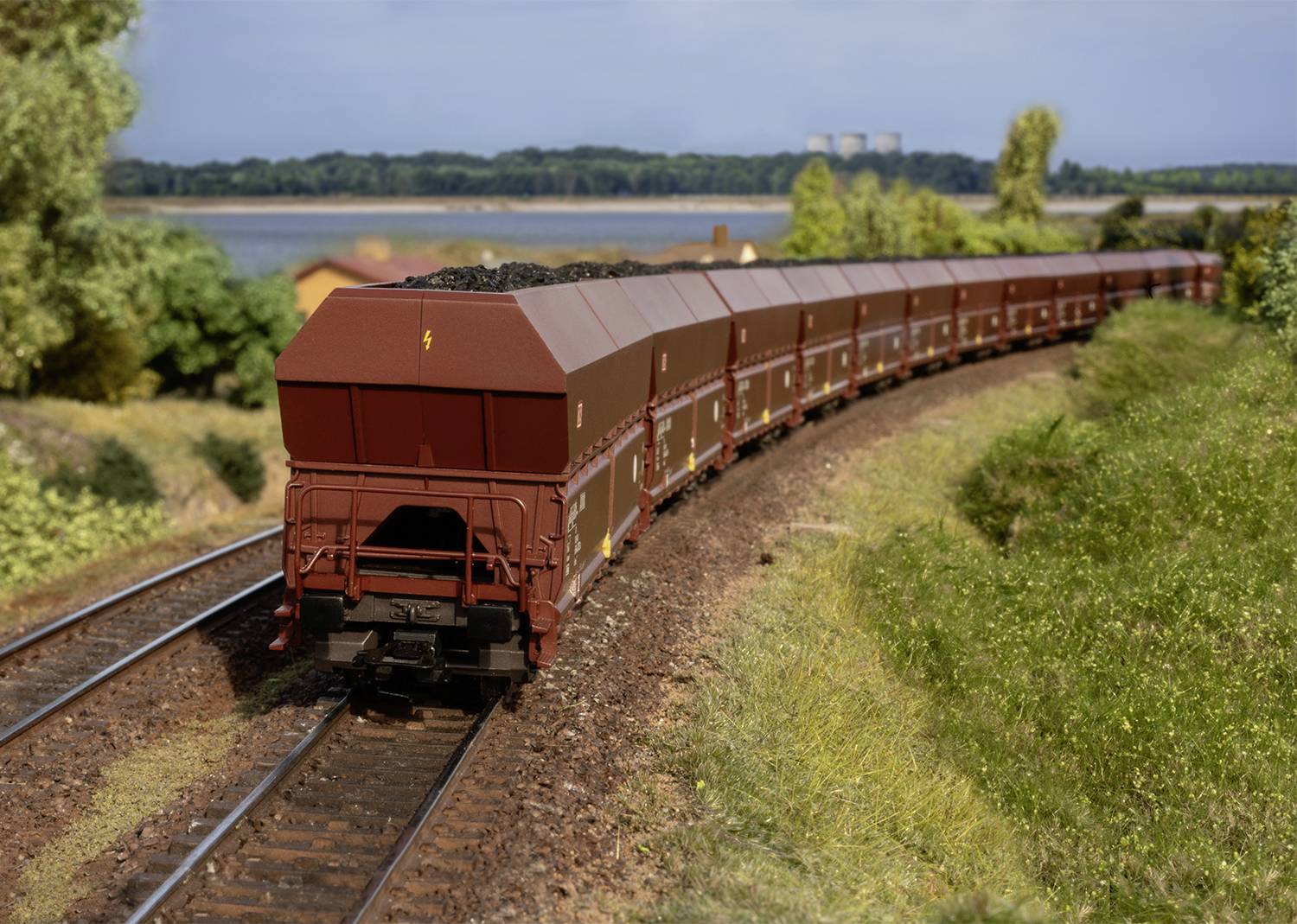 A long freight train with open wagons full of coal travels along a rural route. Trees and a lake can be seen in the background.