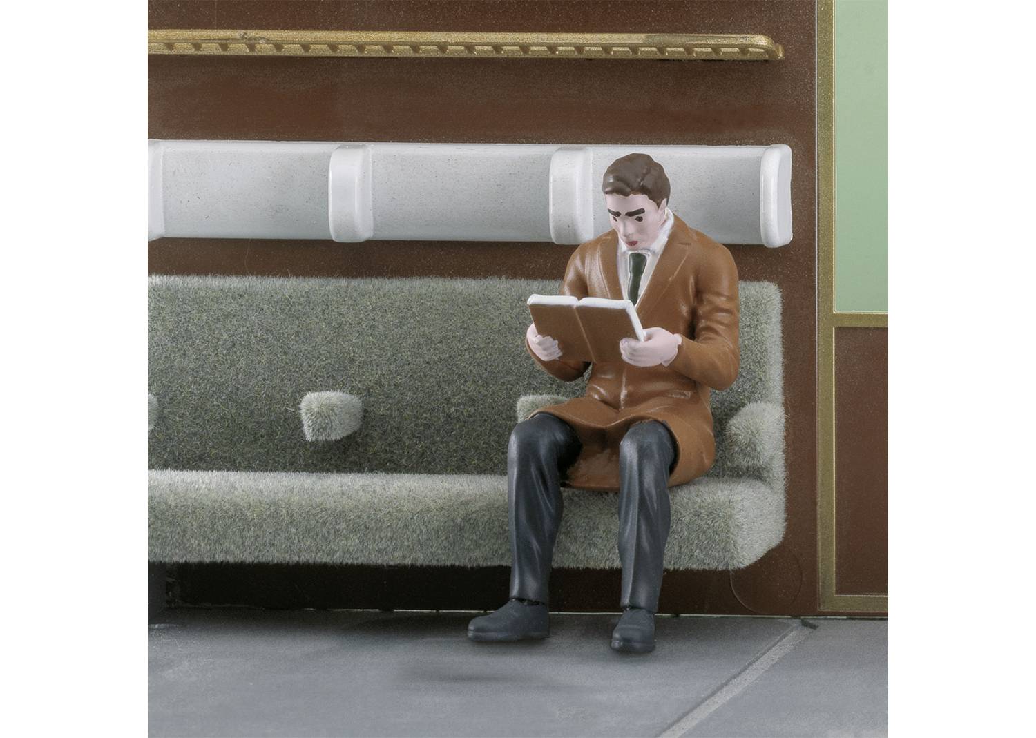 A model figure sits on a bench reading a book. Details of a miniature room model are visible in the background.
