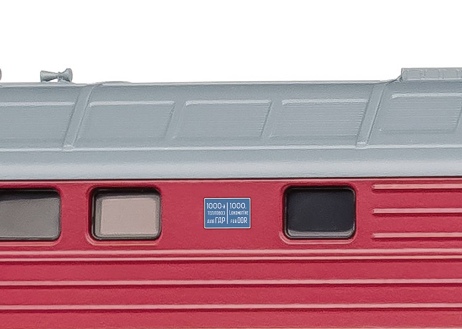 Side view of a red train carriage, part of a railway, with a grey roof and small windows. A blue sign with writing is visible.