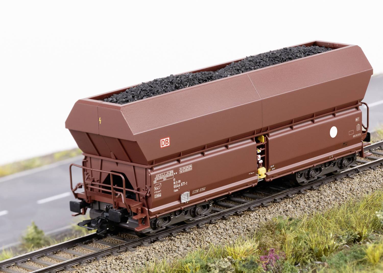 A brown freight wagon of Deutsche Bahn, loaded with coal, stands on railway tracks. A road and grass are visible in the background.