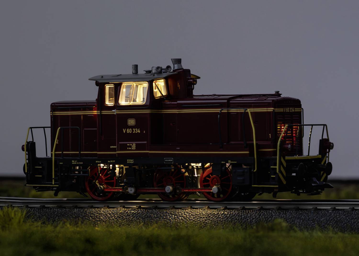 A model railway locomotive in dark red with an illuminated cab travels along tracks, surrounded by grass and a dark background.