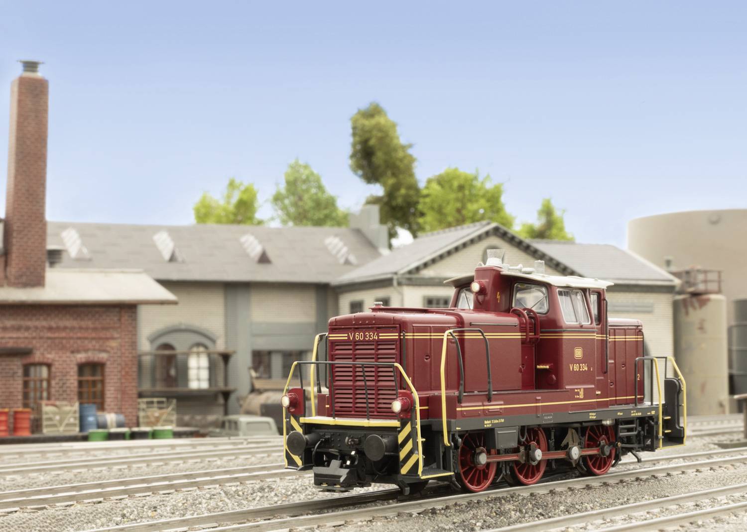 A red model locomotive travels along tracks in front of a background of buildings and trees.