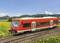 Red train travelling on railway tracks through a green, blooming landscape with hills in the background under a blue sky.
