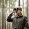 A person in the woods holding a pair of binoculars, seemingly observing. They are wearing a cap and hunting attire. The surroundings show blurred trees.