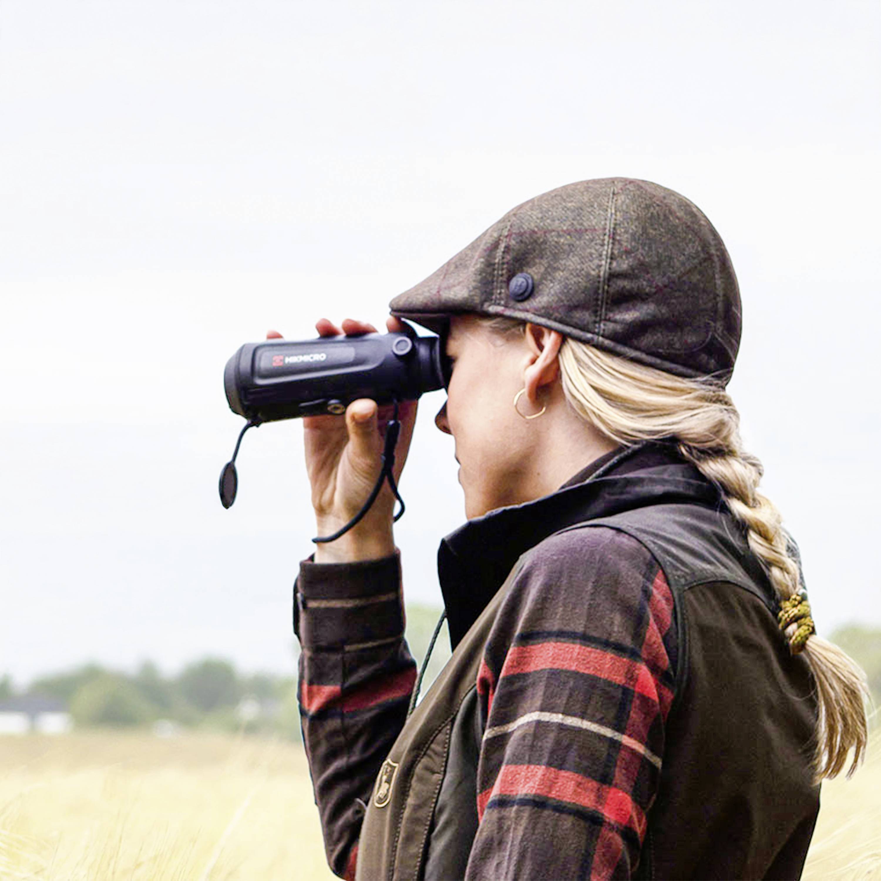 A person is observing a field through binoculars, wearing a checked jacket and a peaked cap.