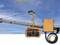 A construction crane against a blue sky. In the foreground, an orange remote control and its cable are positioned, capable of controlling the crane.