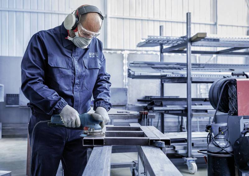 A worker wearing protective clothing is grinding a piece of metal in a factory hall. Metal shelving with various parts can be seen in the background.