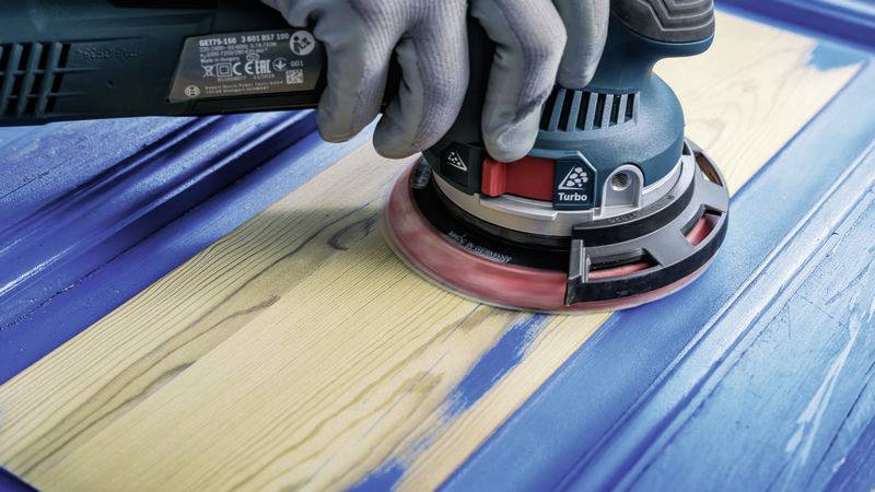 A person is sanding a blue wooden door with an electric sander to prepare the surface for a new coat of paint.