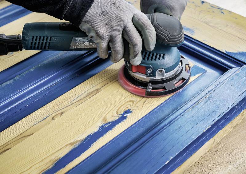 A person is sanding a wooden door painted blue in order to renovate it. The person is wearing gloves and using an electric sander.