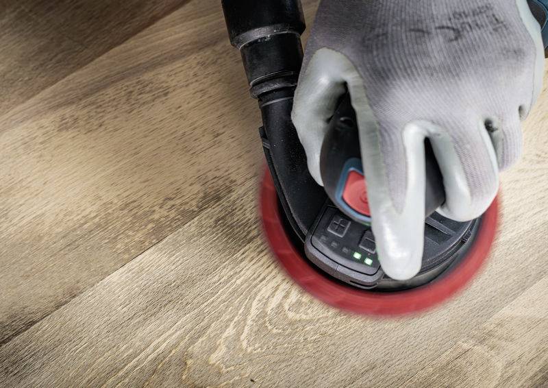 A person is sanding wood with an orbital sander. The sander has a red sanding paper and is being operated by a gloved hand.
