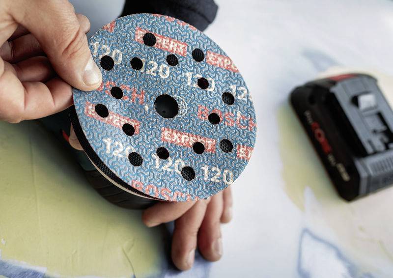 A hand is holding a round piece of sandpaper with holes, clearly intended for an electric tool. A device component is blurrily visible in the background.
