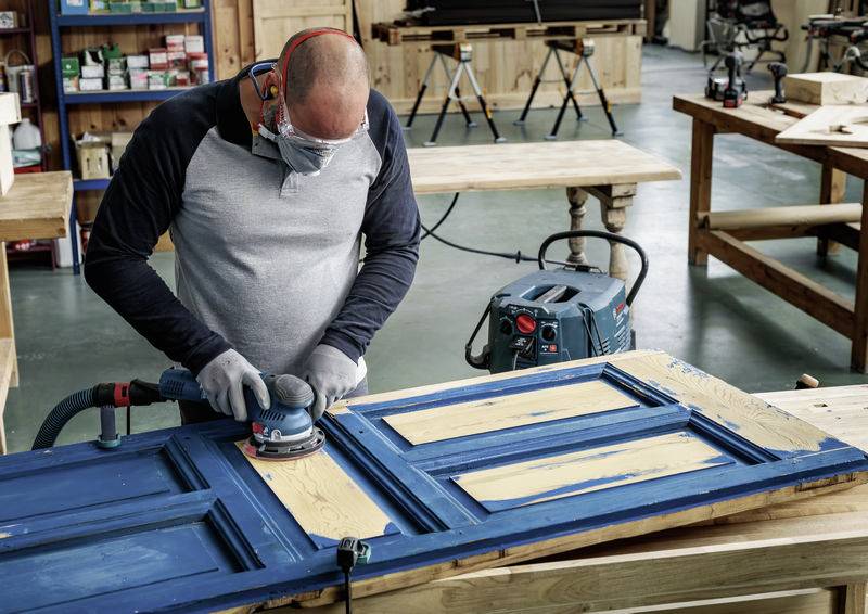 A man is sanding a blue wooden door in a workshop. He is wearing safety glasses and ear protection. Workbenches and shelves are visible in the background.
