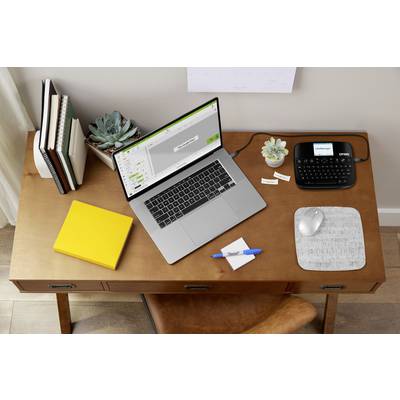 A laptop and label printer on a desk with plants, a notepad and pen, showing an organised workspace.