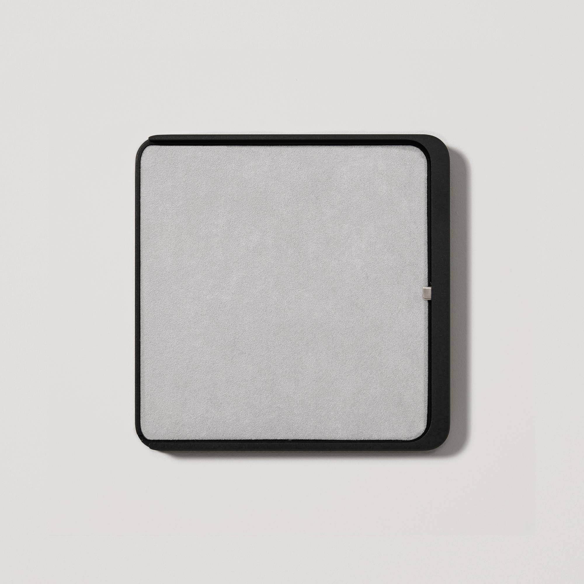 A square, black air purification device with a grey filter insert, mounted on a white wall.