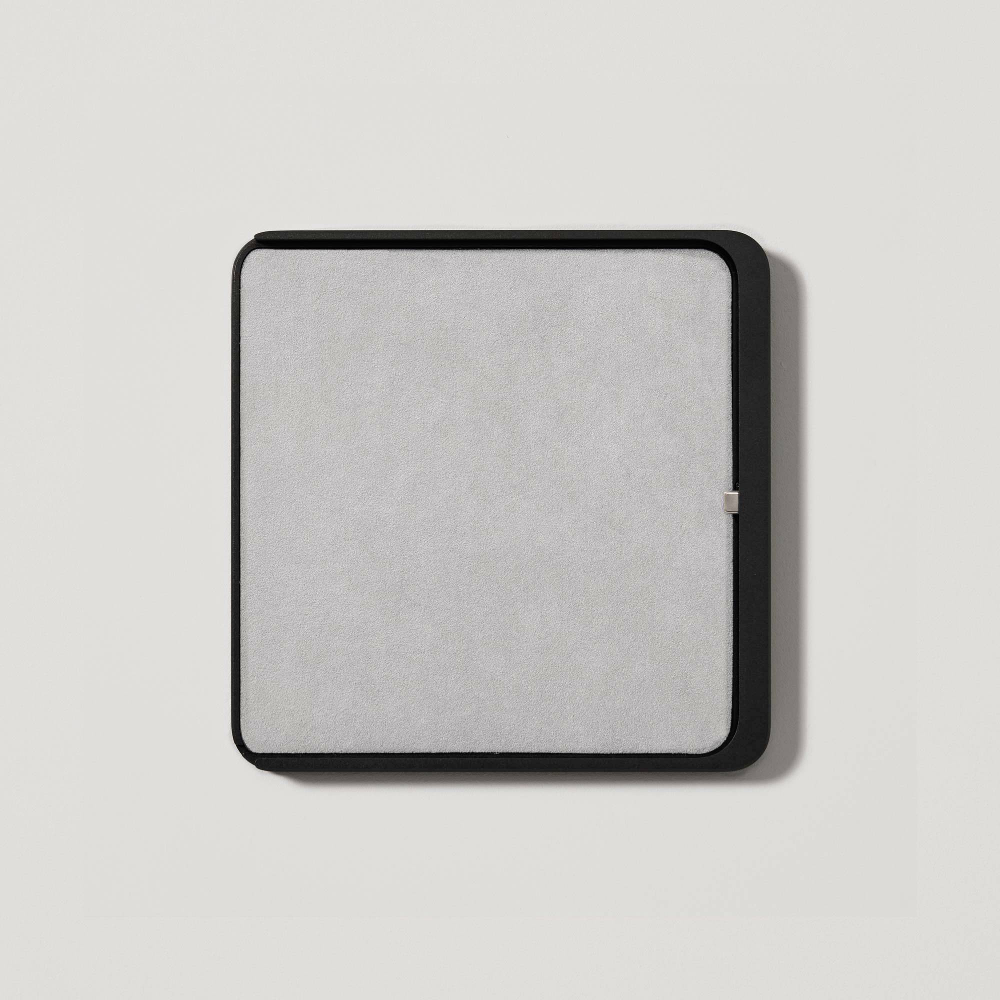 A square air purifier with a black frame and light grey fabric surface, mounted on a light-coloured wall.