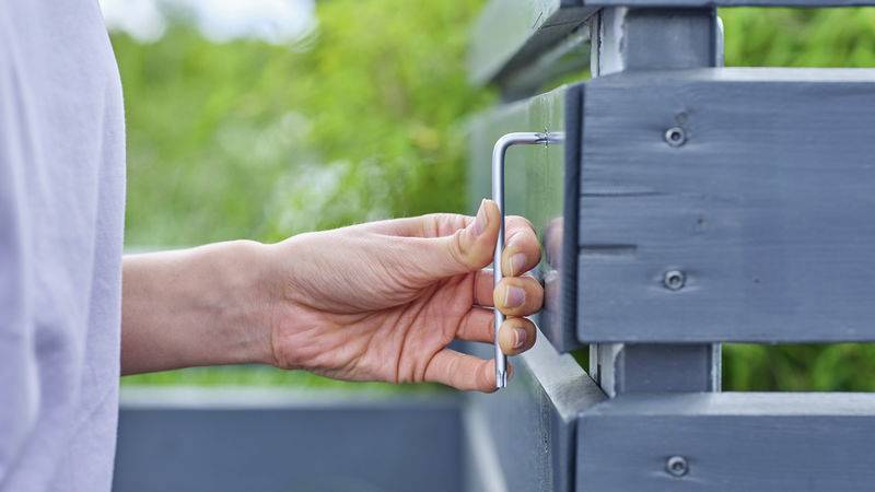 A hand is turning an Allen key to secure a light grey wooden structure outdoors. Green scenery is visible in the background.