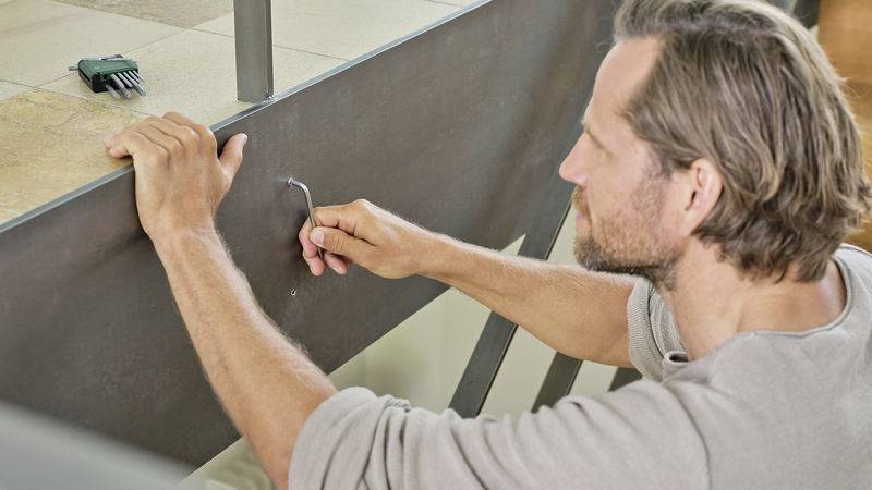 A man is securing a metal plate with an Allen key. A tool lies on the ground in the background.