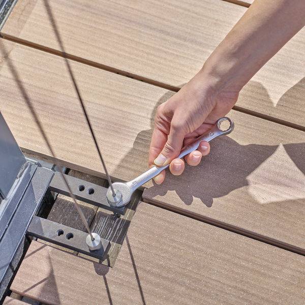 A hand tightens a screw with a spanner on a metal clamp on a wooden floor.