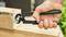 A hand holds a pair of pliers and pulls a nail from a wooden board. The pliers have green handles.