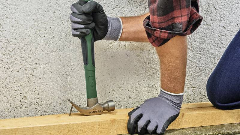 A person wearing gloves is hammering a nail into a wooden board lying on the ground.