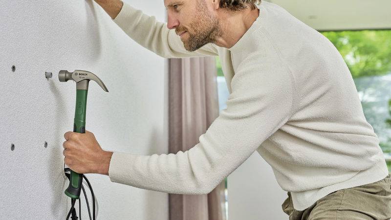 A man hammers a nail into a white wall. He is wearing a white top and is focused on his work.