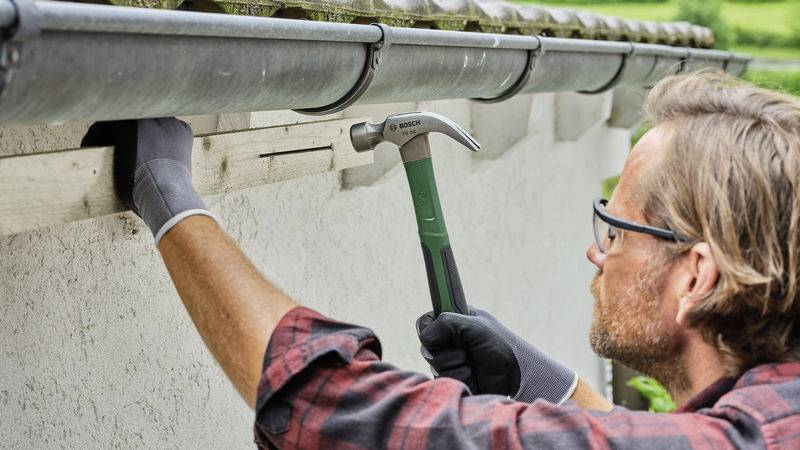 A man is repairing the guttering of a house with a hammer, wearing gloves and a shirt.