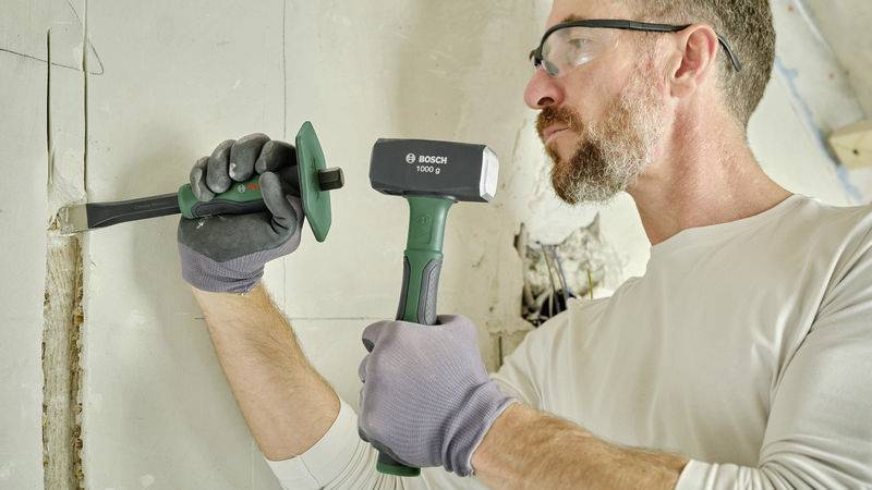 A man wearing safety glasses and gloves is working with a hammer and chisel on a wall in a dilapidated room.