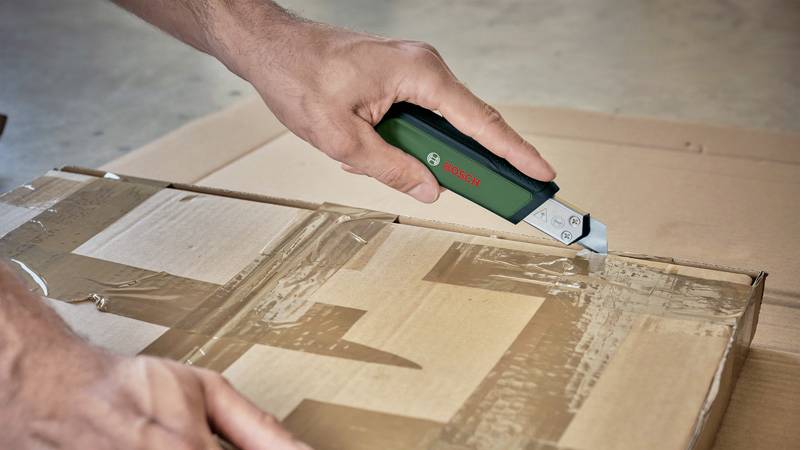 A person is using a green craft knife to open a package sealed with adhesive tape.