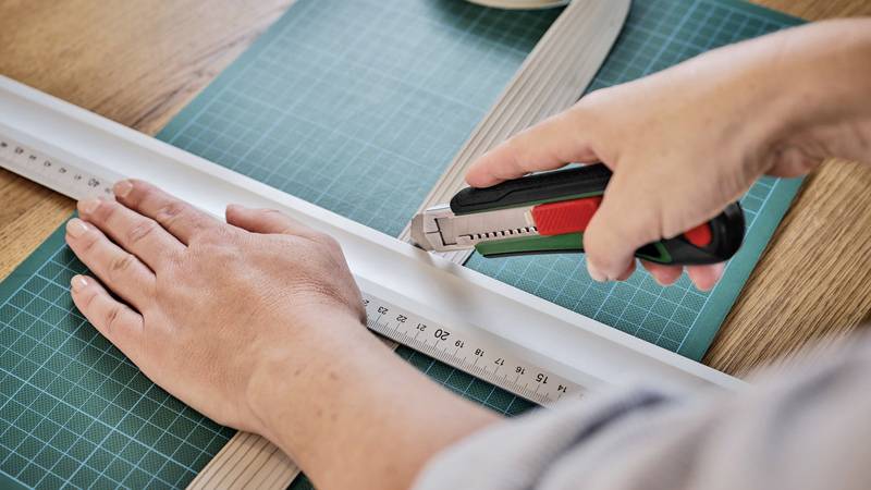 A person is cutting a piece of plastic with a craft knife along a ruler on a green cutting mat.