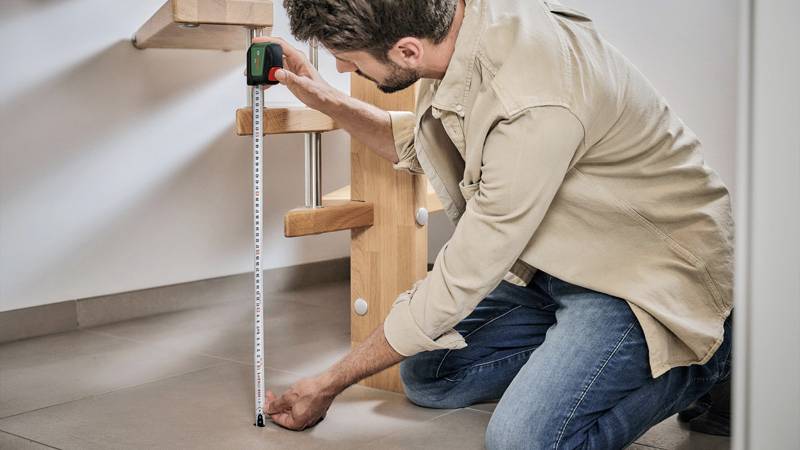 A man is kneeling on the floor and measuring the height of a wooden staircase with a tape measure in a flat.