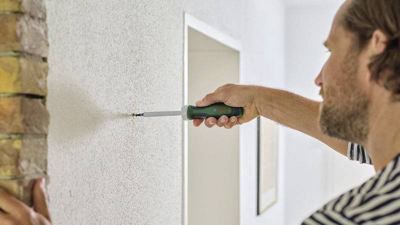 A man is using a screwdriver to twist a screw into a white wall. Bricks are visible on the left side of the image.